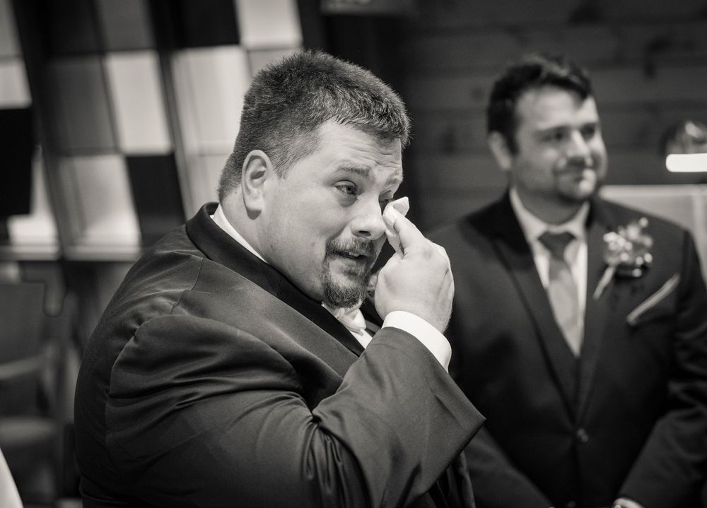 Emotional groom wiping tears during wedding ceremony in black and white photo.