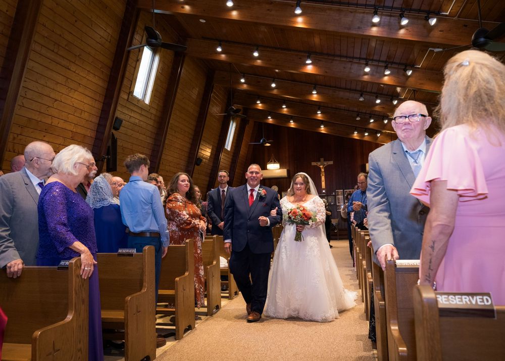 Bride and groom walk down the aisle of a wooden church chapel surrounded by seated wedding guests.