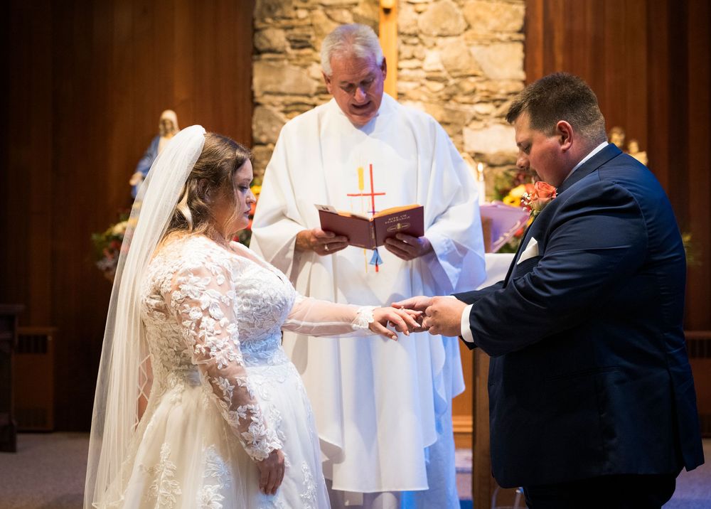 Bride and groom exchange rings during wedding ceremony with officiant holding red book at church altar.