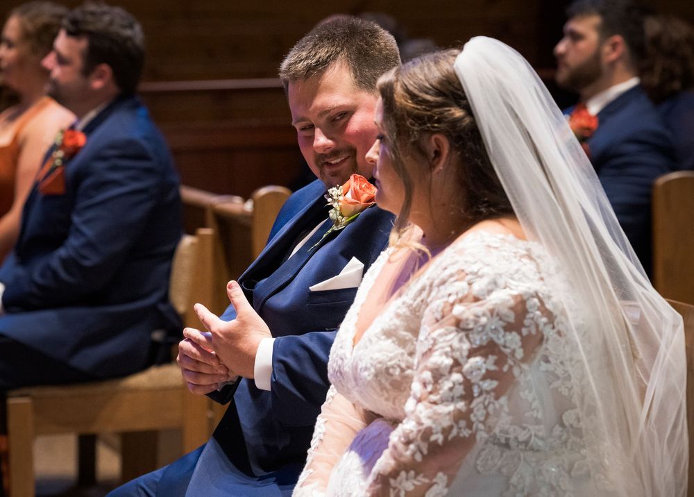 Bride and groom smiling joyfully during wedding ceremony in church, bride wearing lace gown and veil, groom in navy suit.