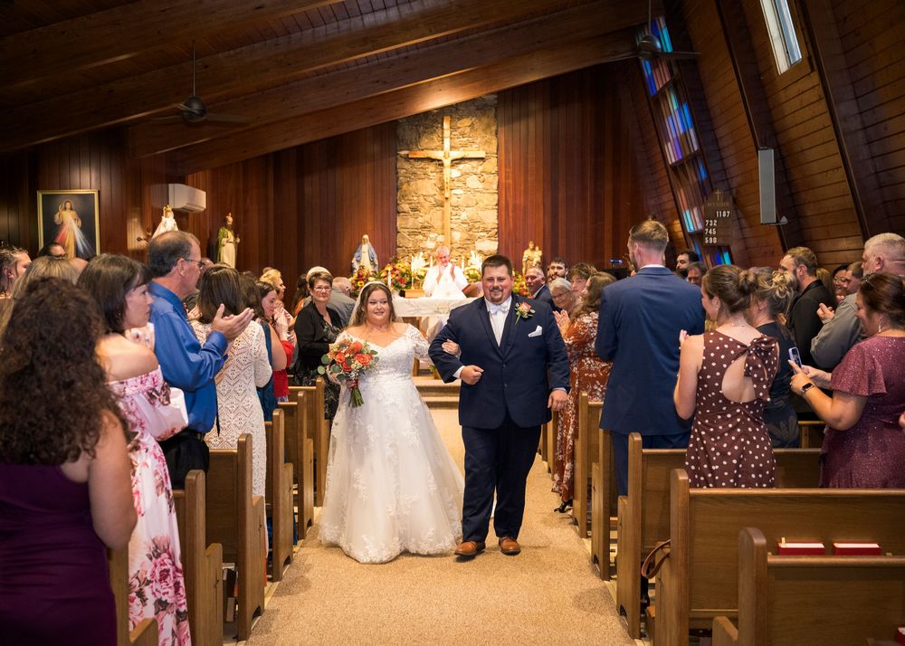 Bride and groom walk down church aisle after wedding ceremony, guests applauding in wooden pew chapel.