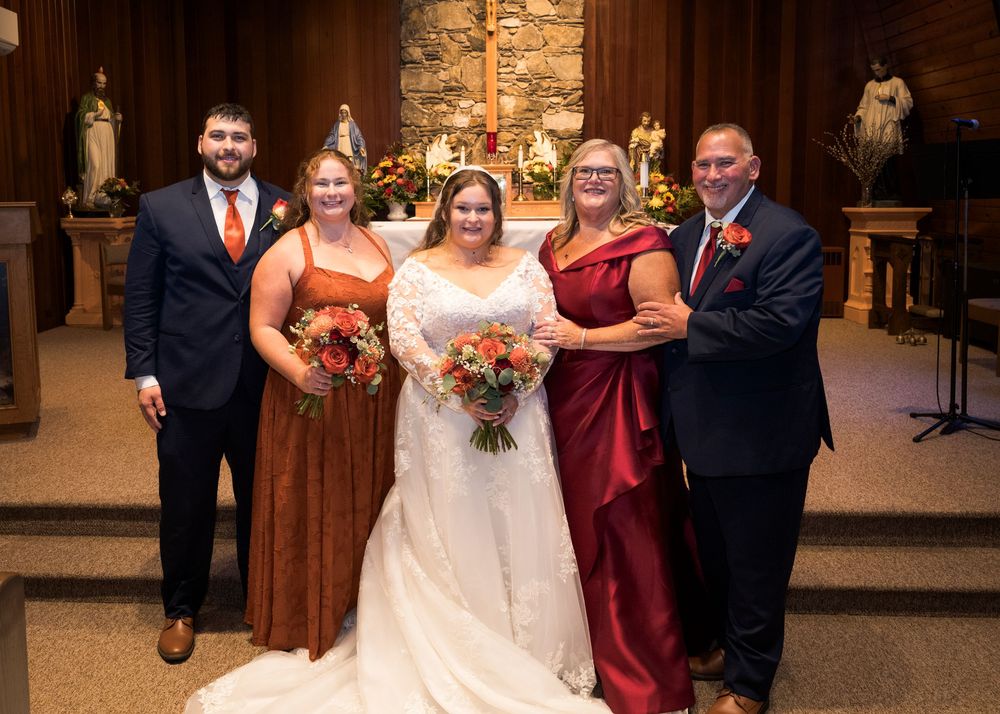 Bride in white gown poses with family in church, women in rust and red dresses, man in navy suit, floral bouquets.