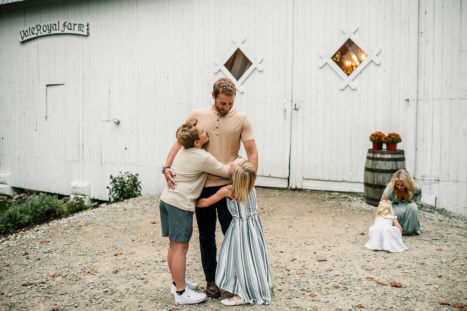 Family embracing outside a white barn at Vale Royal Farm, with a young girl in a blue dress nearby.