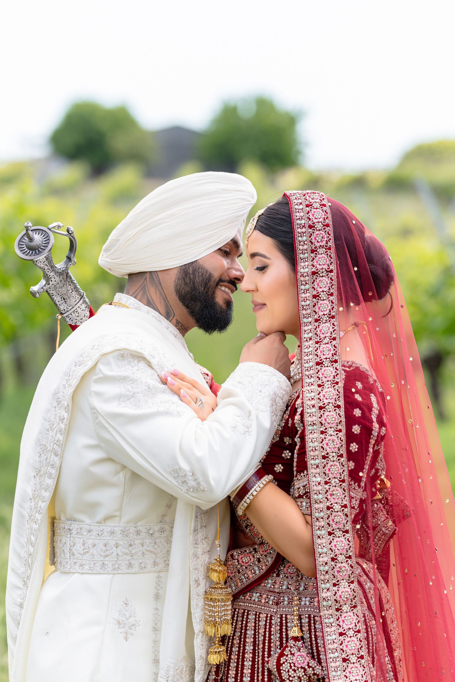 A Sikh groom in white sherwani and turban gazes lovingly at his bride in red velvet lehenga and dupatta outdoors.