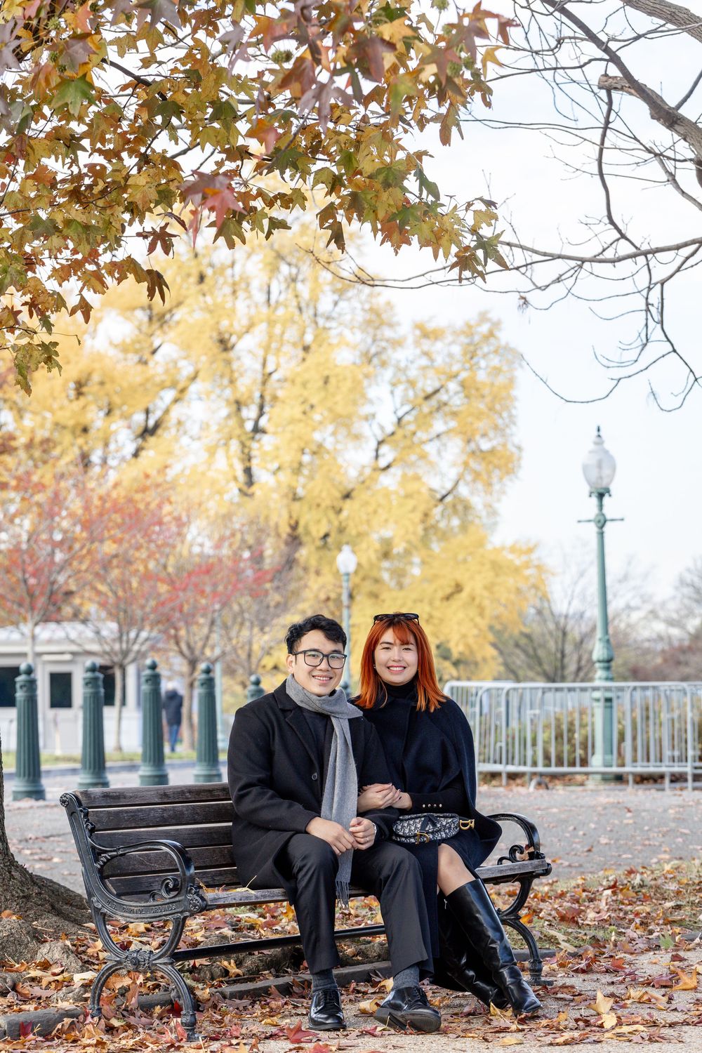 Couple sitting on a park bench surrounded by colorful autumn foliage, smiling together on a fall day.