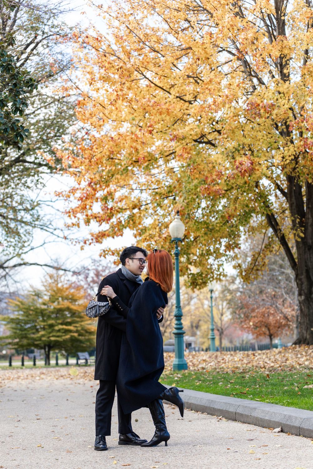 Couple embracing on a park path surrounded by vibrant autumn foliage during fall season.