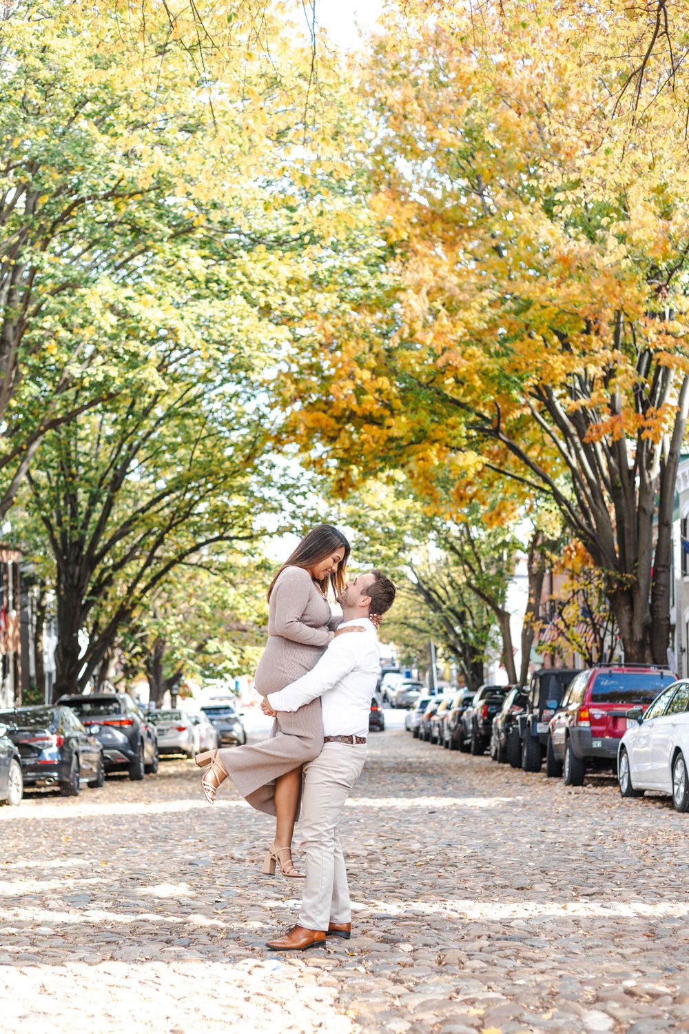 Couple embracing on a cobblestone street lined with autumn trees and parked cars in fall.