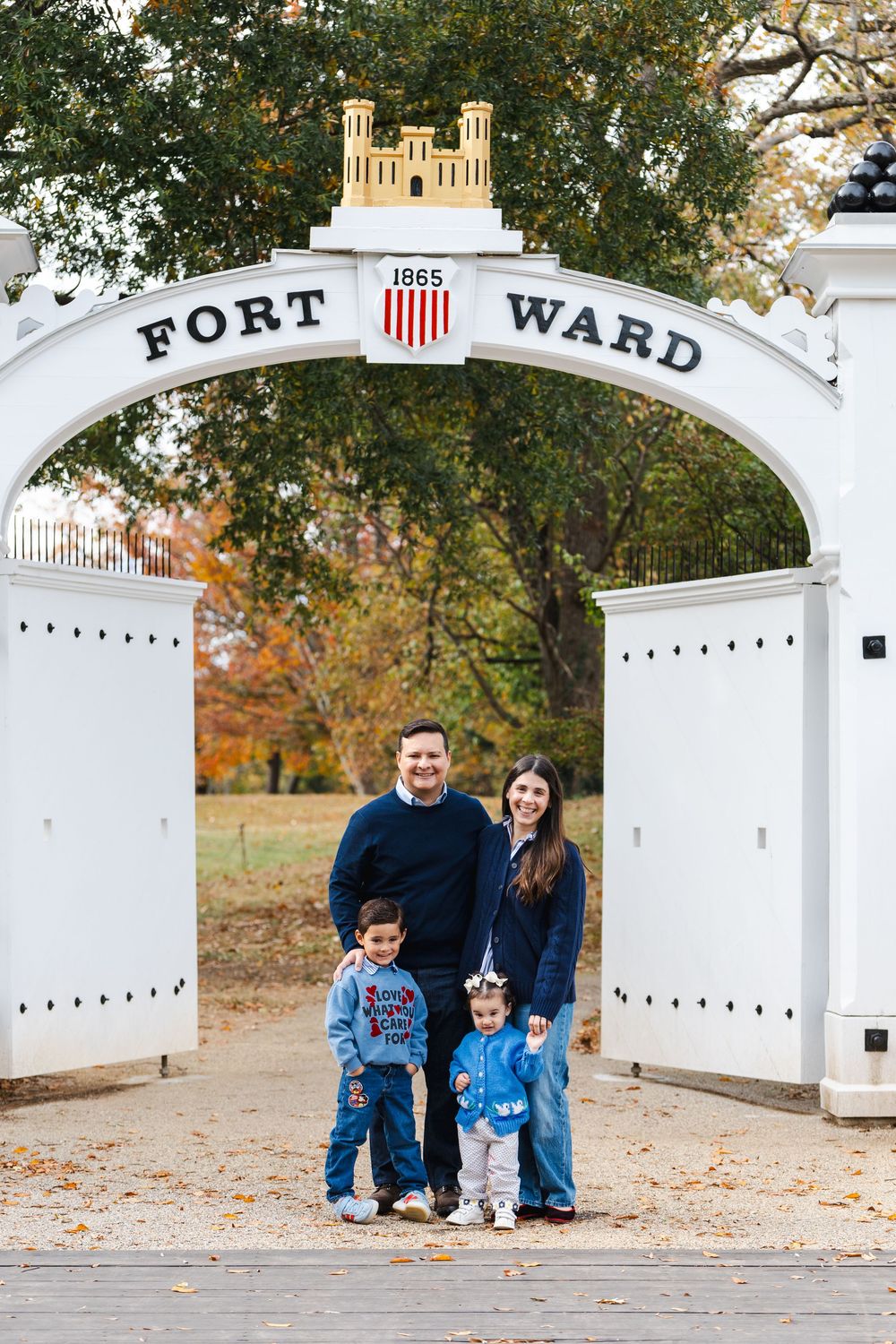 Family of four posing under the historic Fort Ward arch entrance, surrounded by autumn foliage in Alexandria, VA.