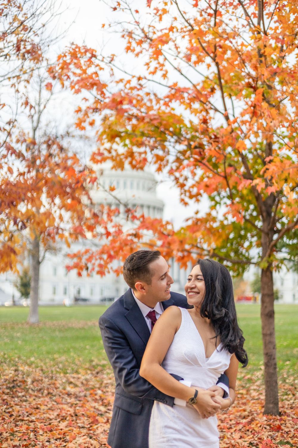 Couple embracing in autumn park with colorful orange fall foliage and US Capitol building in background.