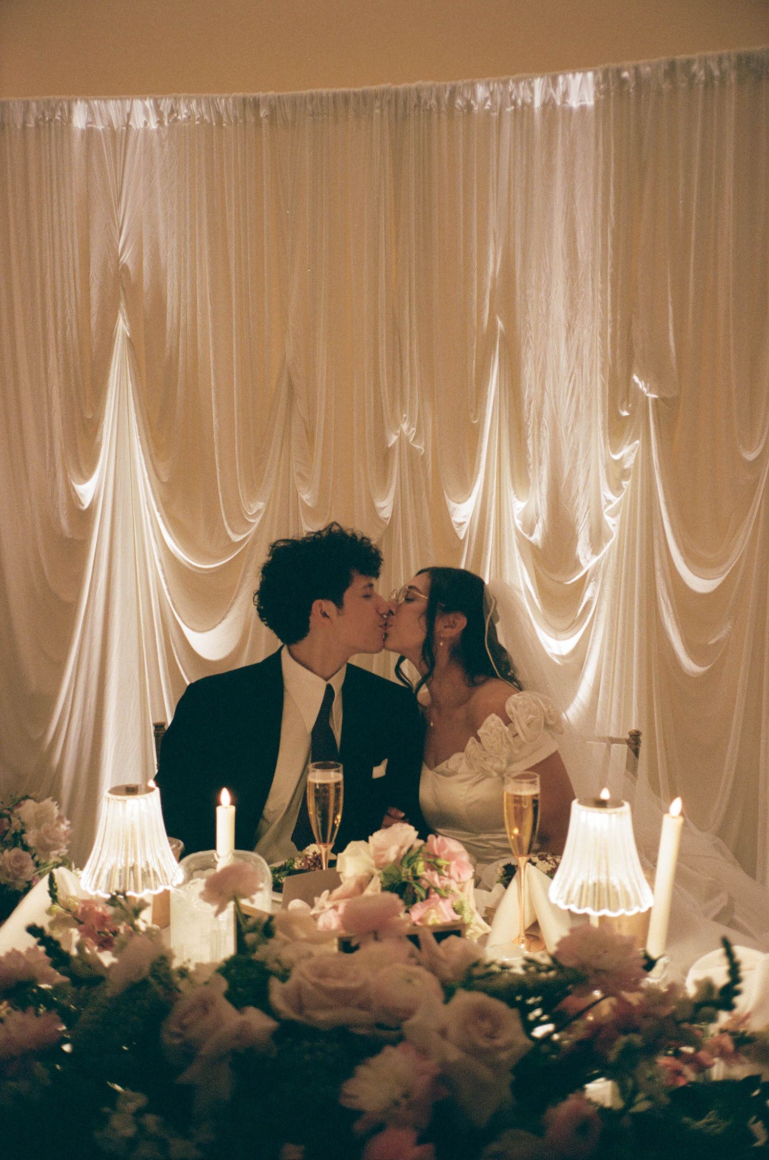 A bride and groom share a kiss at their wedding reception table adorned with roses and candles before a draped white backdrop.