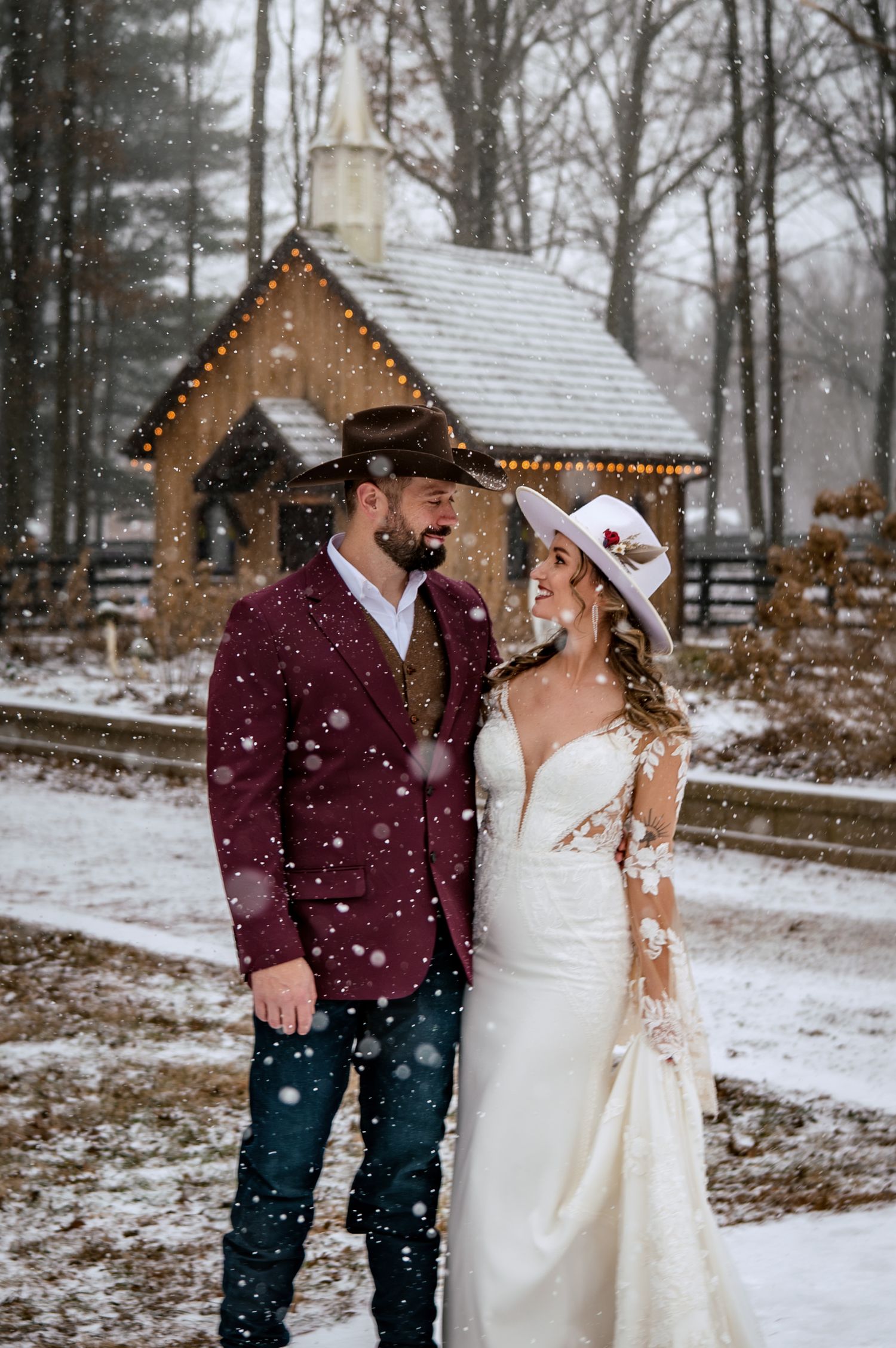 Couple in western attire poses in snow before rustic chapel, groom in burgundy blazer and bride in lace wedding gown.