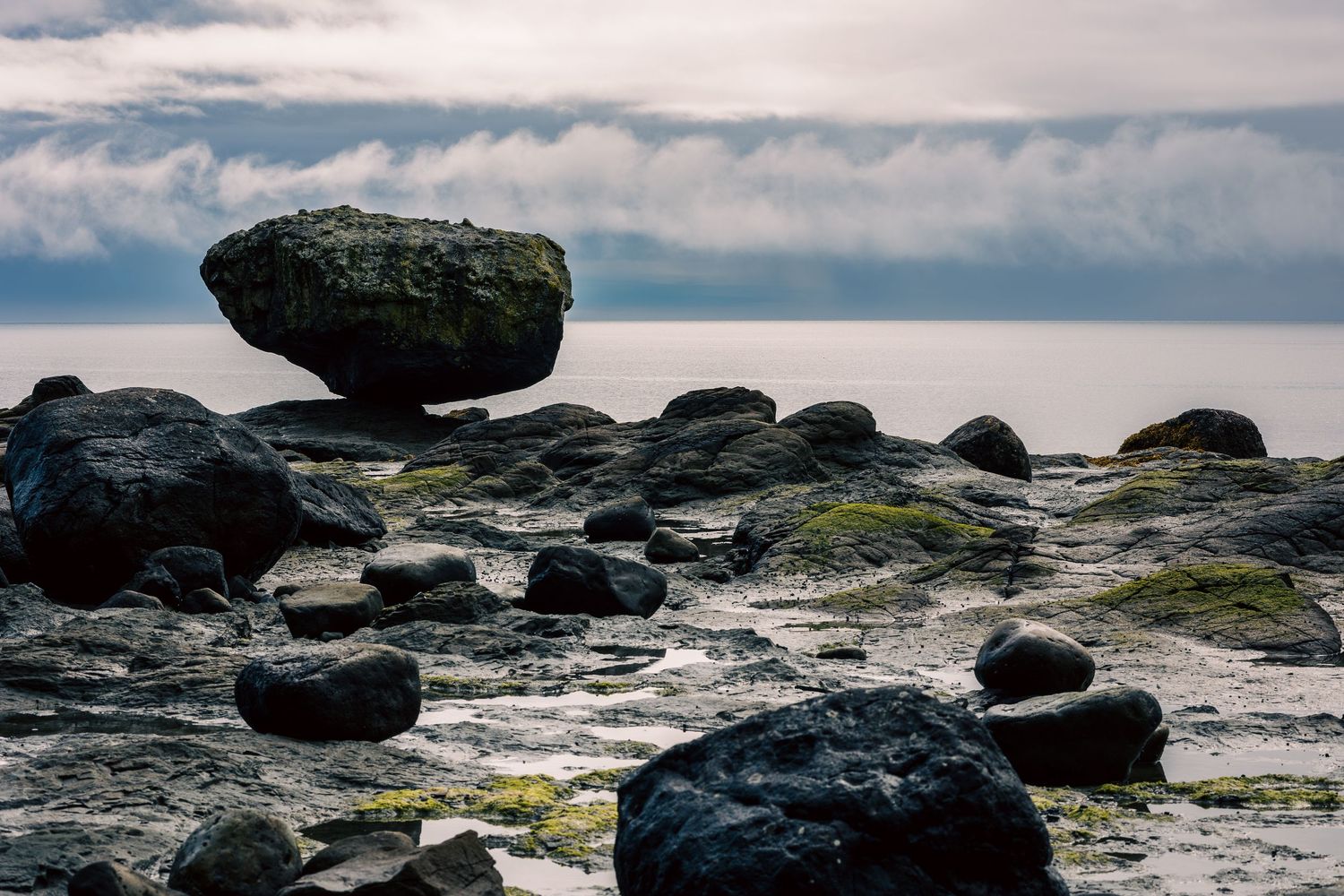 Rocky shoreline with a large balanced boulder perched on mossy rocks, overlooking a calm sea under cloudy skies.