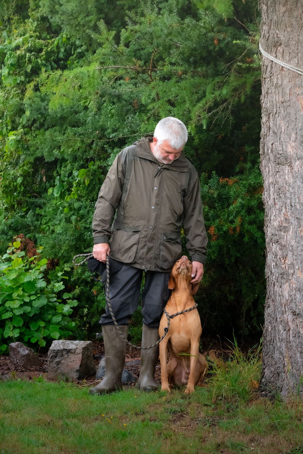 Older man in green jacket and boots stands with a Vizsla dog on a leash in a lush garden setting.