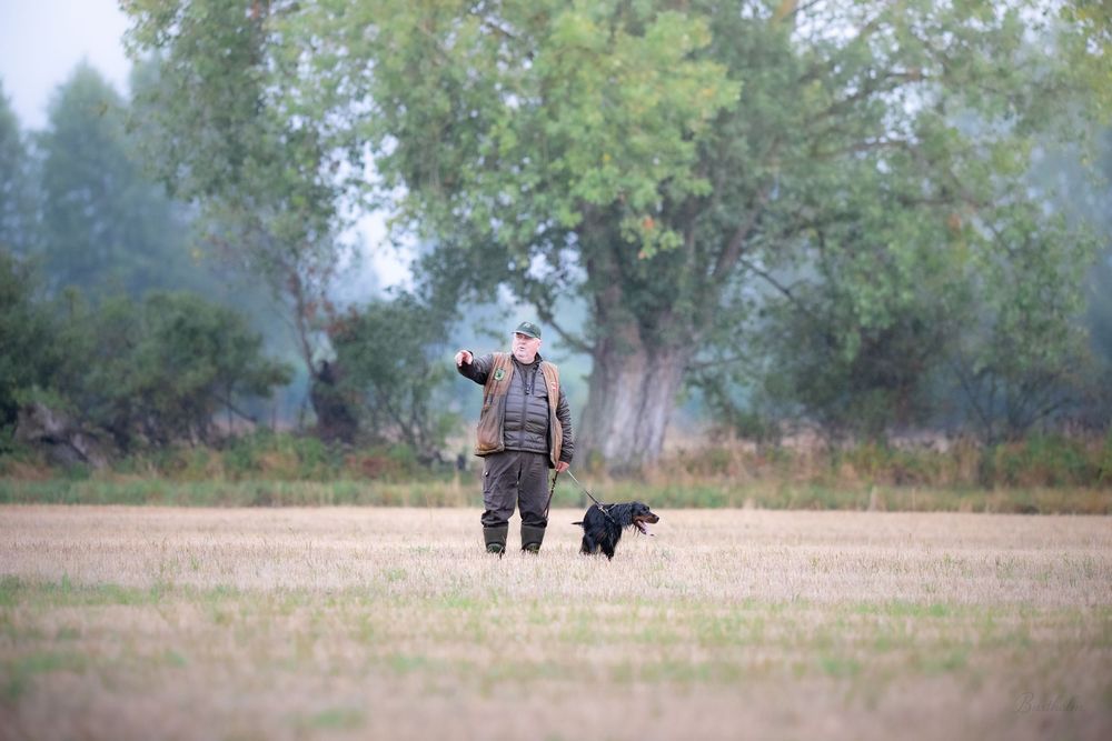 Hunter walking through a field with a black Labrador retriever dog during an outdoor hunting expedition.