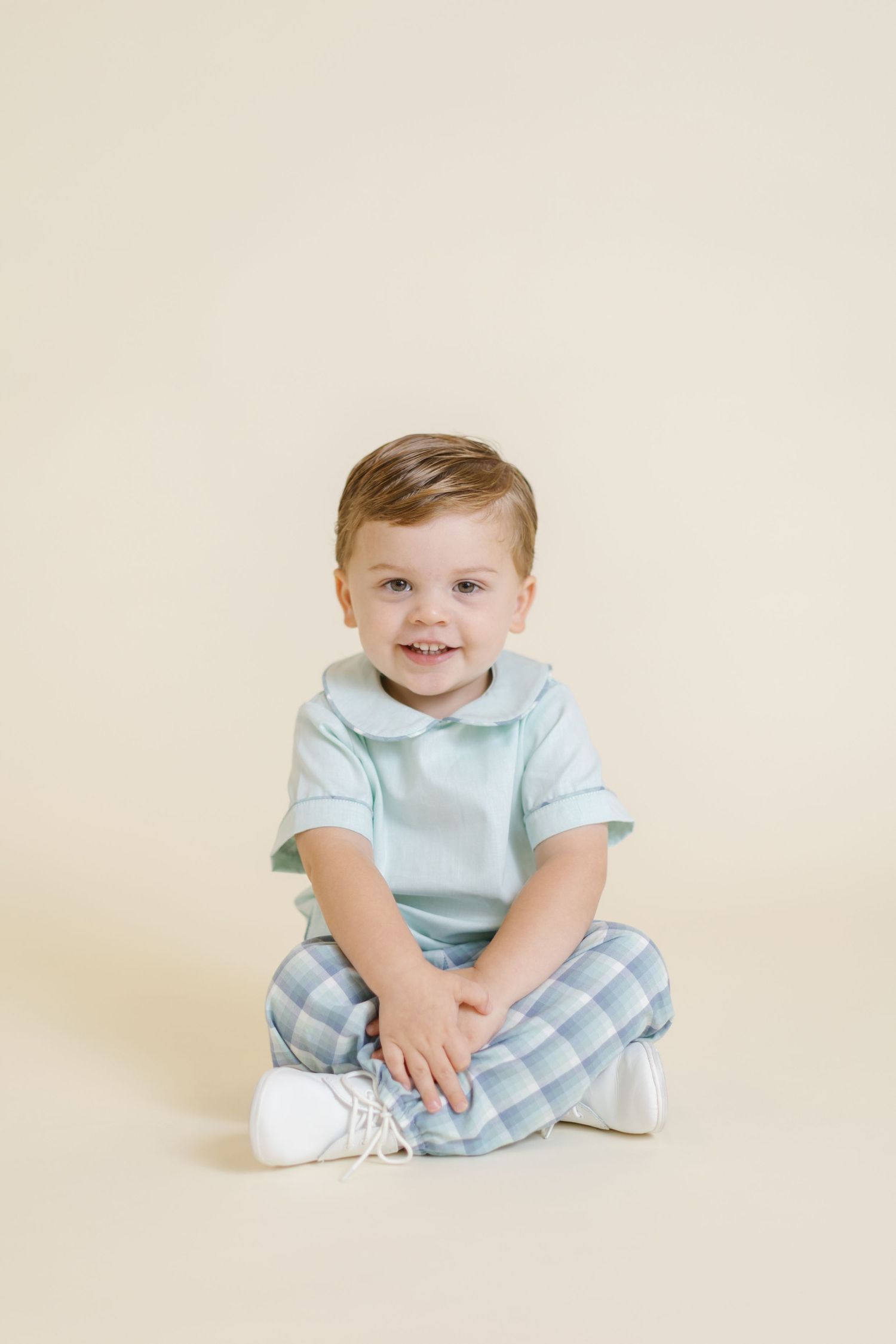 Smiling toddler boy sitting cross-legged in mint top and plaid pants with white shoes on cream background.