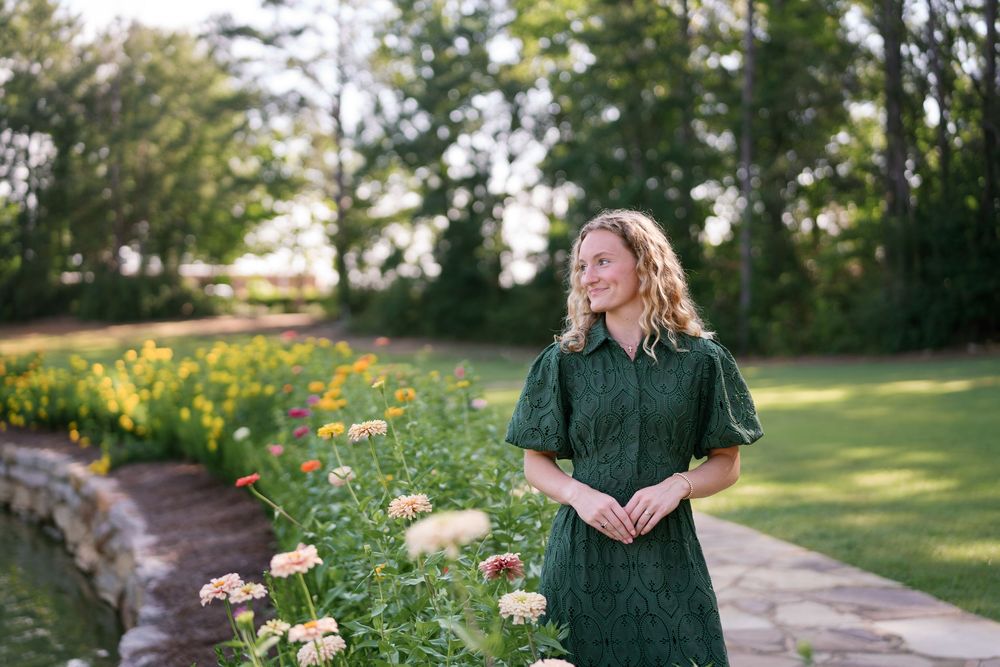 Woman in green dress standing by colorful flower garden with lush trees in background on sunny day.