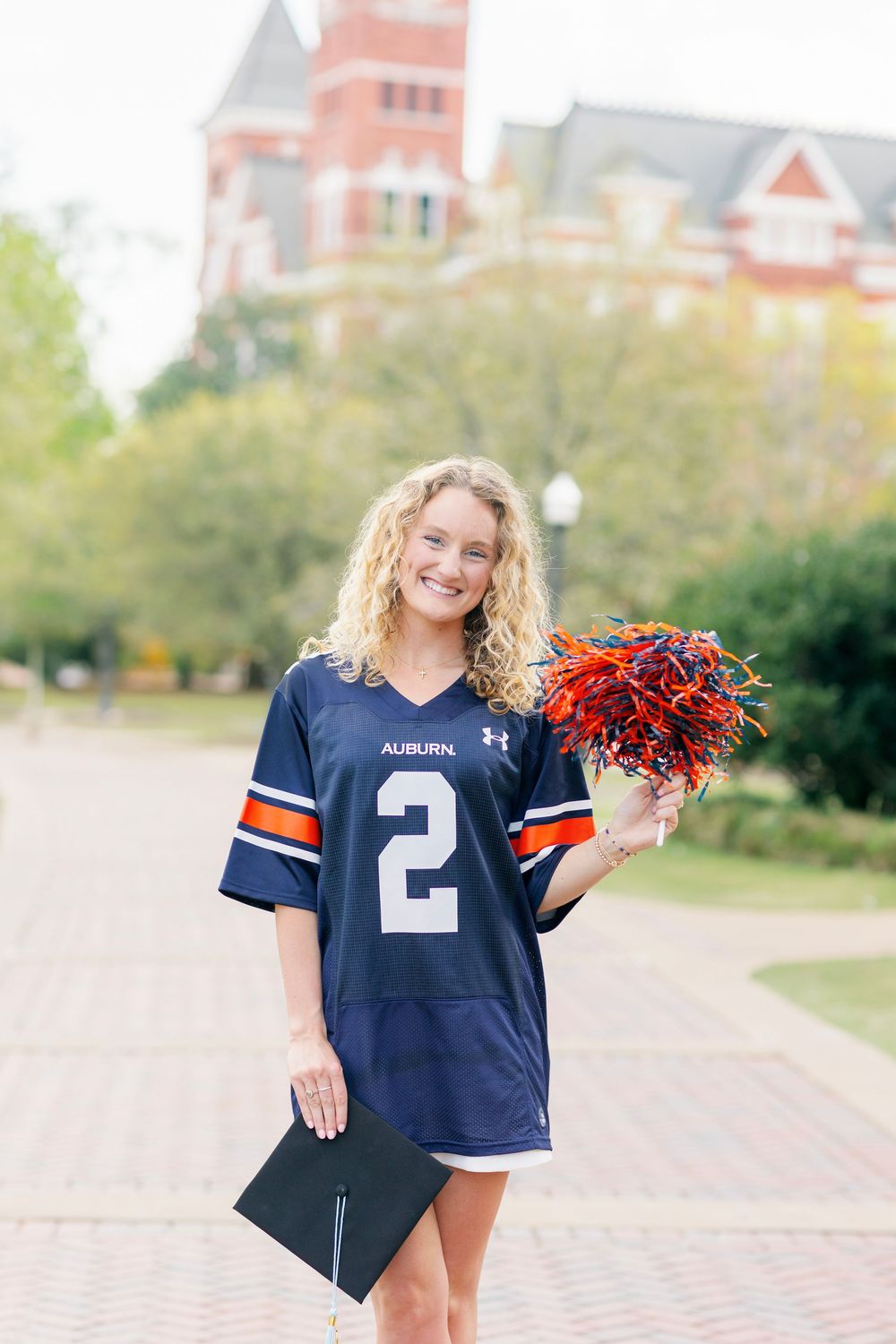 Person in navy football jersey poses with red pom poms against campus backdrop.