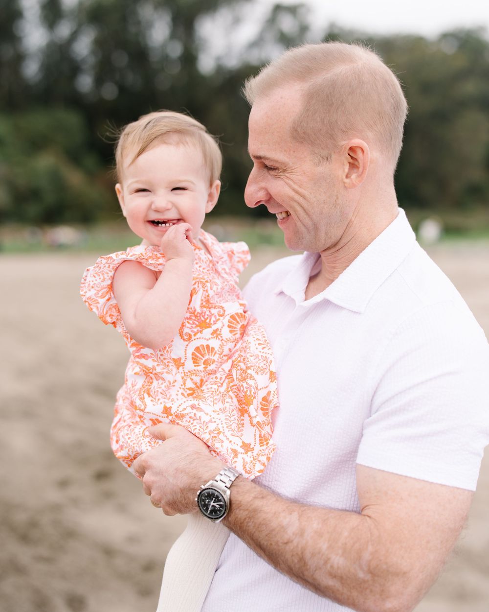 A smiling baby girl in an orange floral romper being held by a man in a white polo on a beach.
