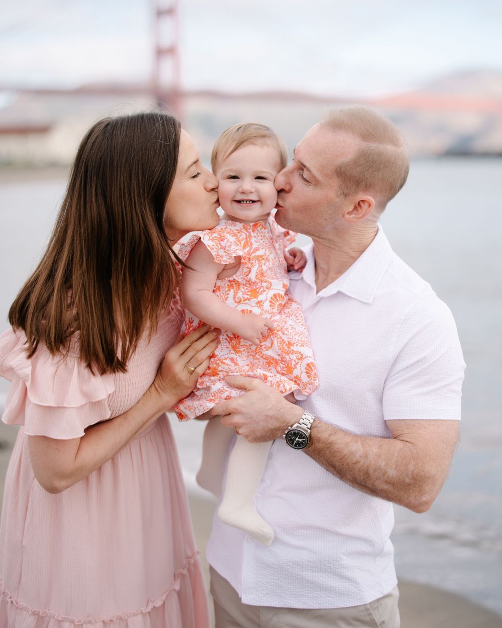 Family photo with parents kissing baby girl by Golden Gate Bridge.
