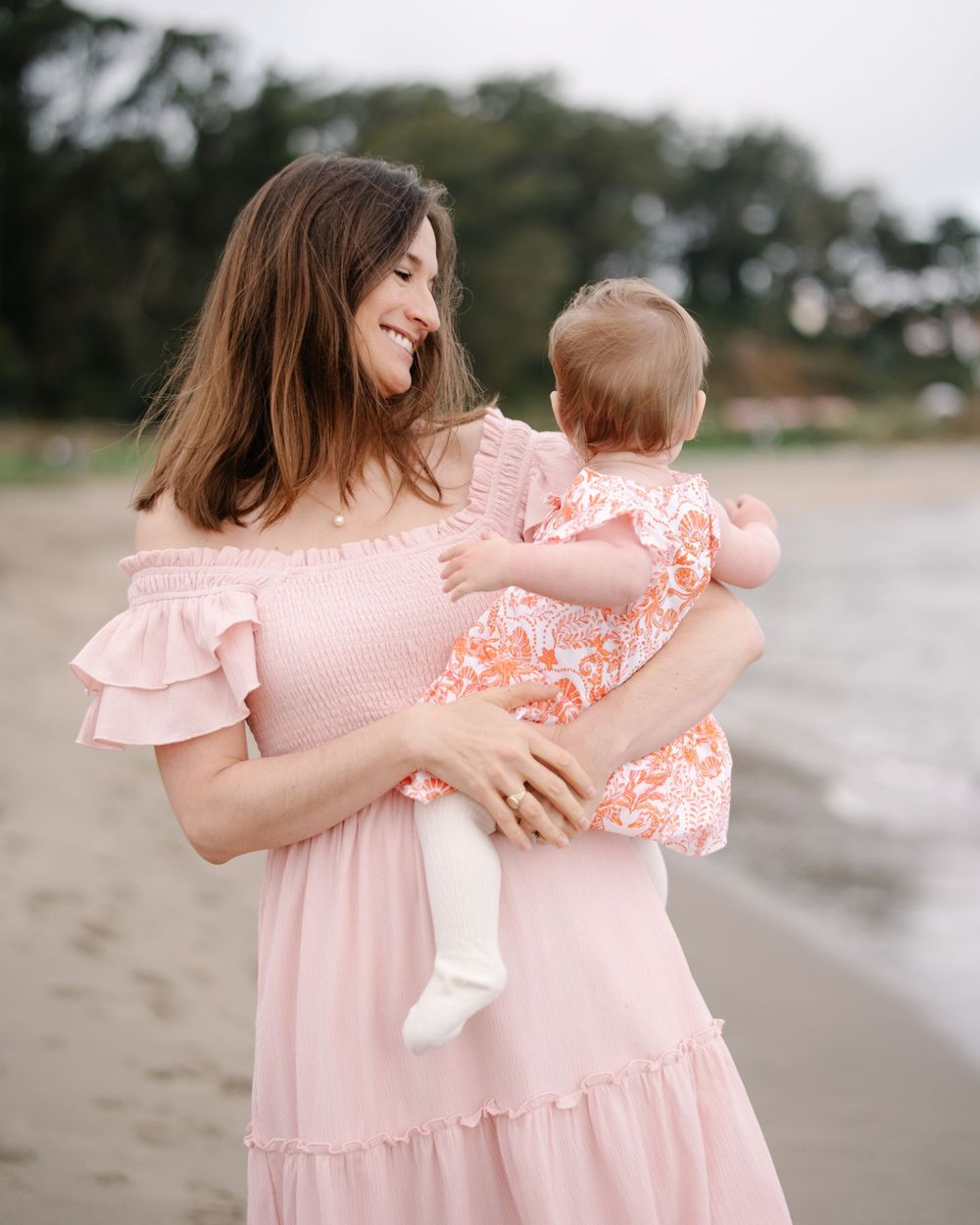 A smiling woman in a pink off-shoulder dress holds her baby girl on a sandy beach.
