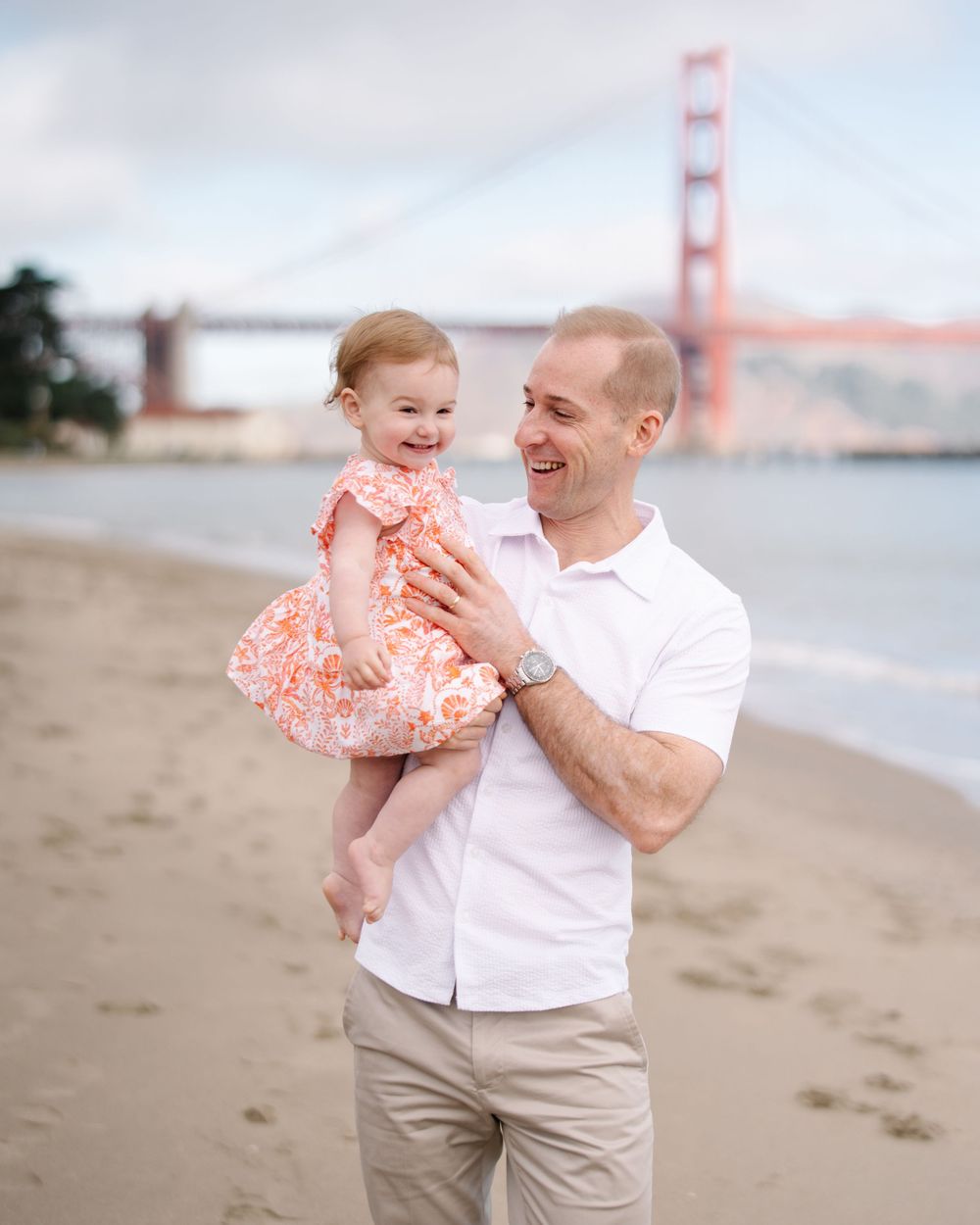 Man holding toddler girl on beach with Golden Gate Bridge in background.