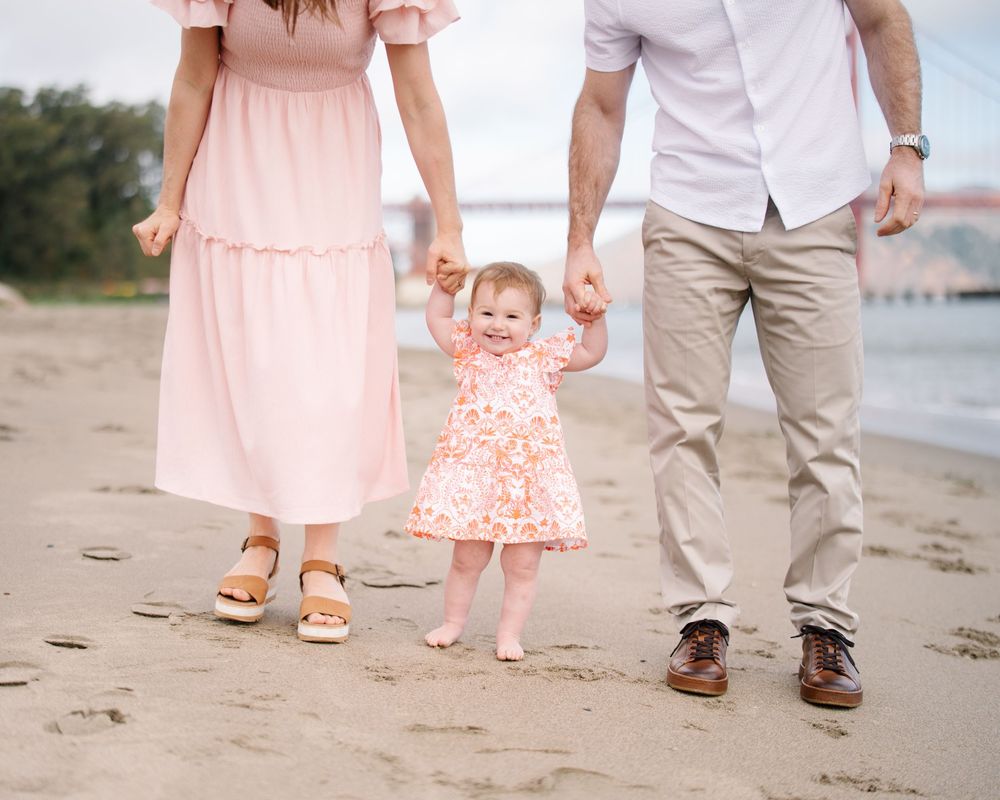 A family of three walking on a sandy beach, parents holding their toddler daughter's hands near a bridge.