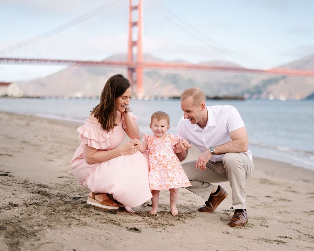 Family with baby girl plays on sandy beach with Golden Gate Bridge backdrop.