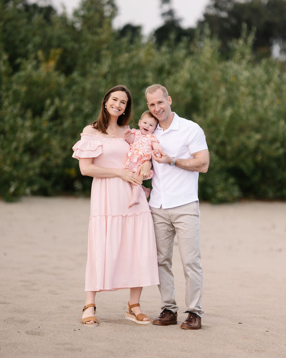 Family of three posing on sandy beach, mother in pink dress holding infant, father in white polo and khaki pants.