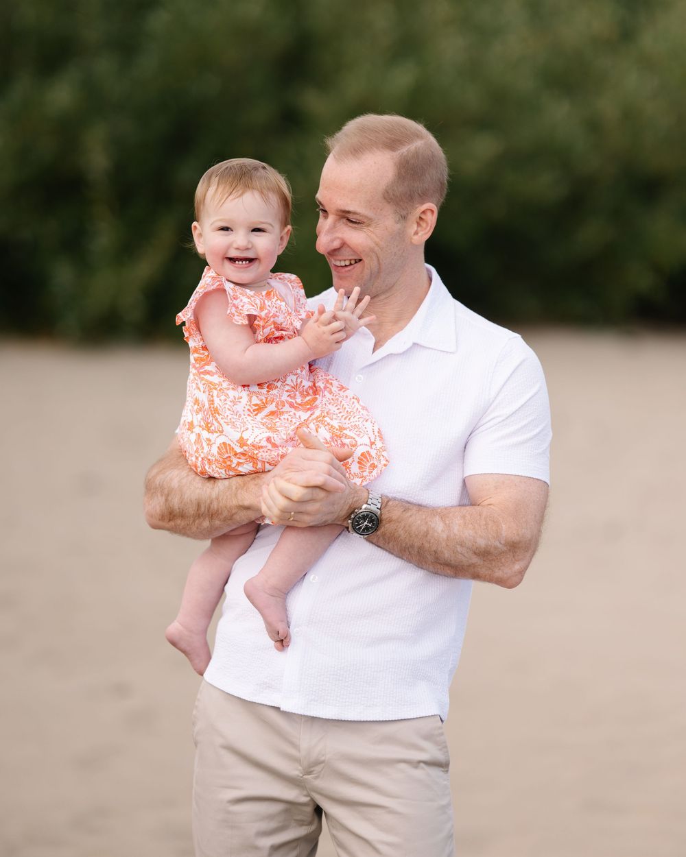A smiling man in a white polo shirt holds a laughing baby girl in an orange floral dress on a sandy beach.