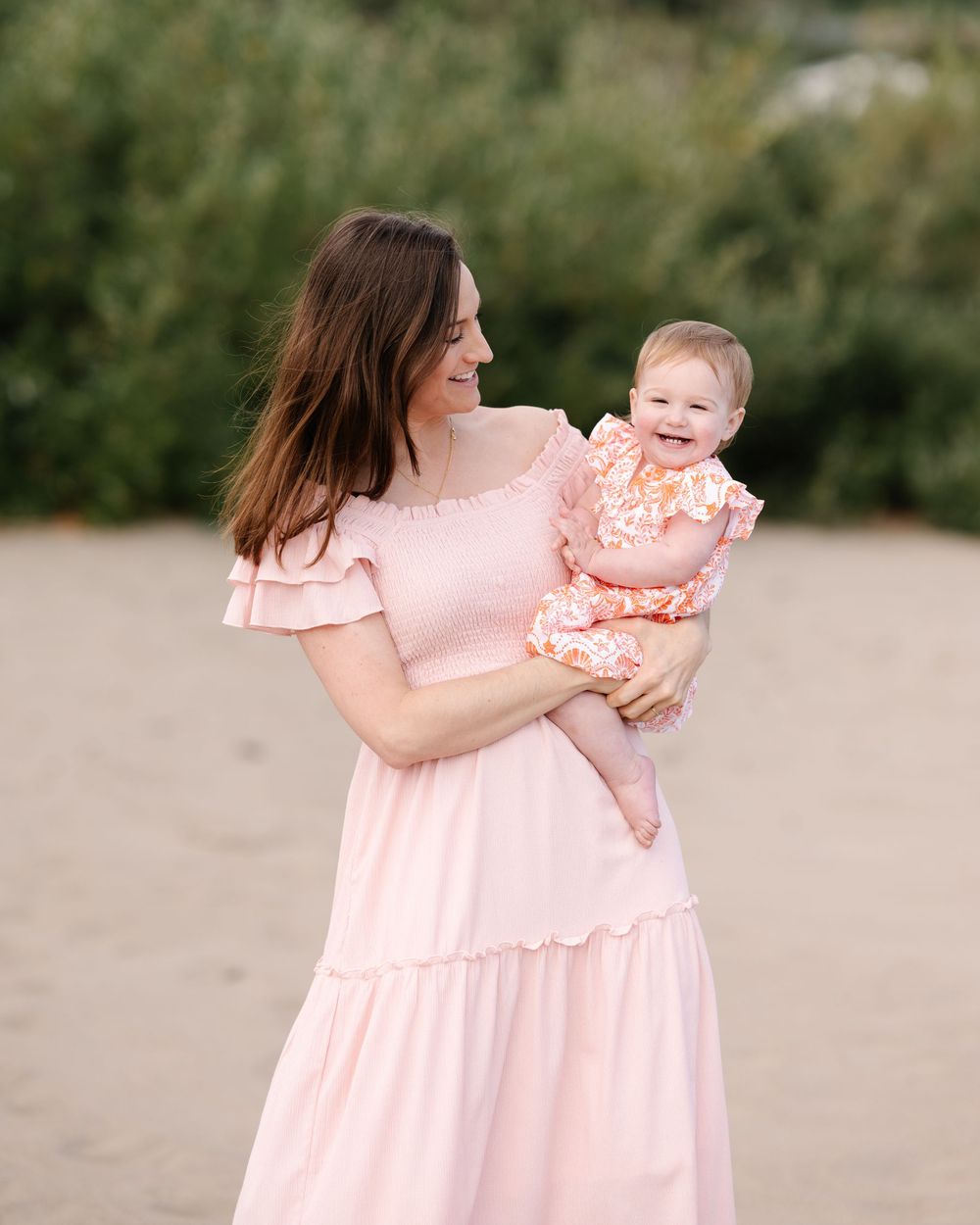 Woman in pink dress holds smiling baby in floral outfit on sandy beach.