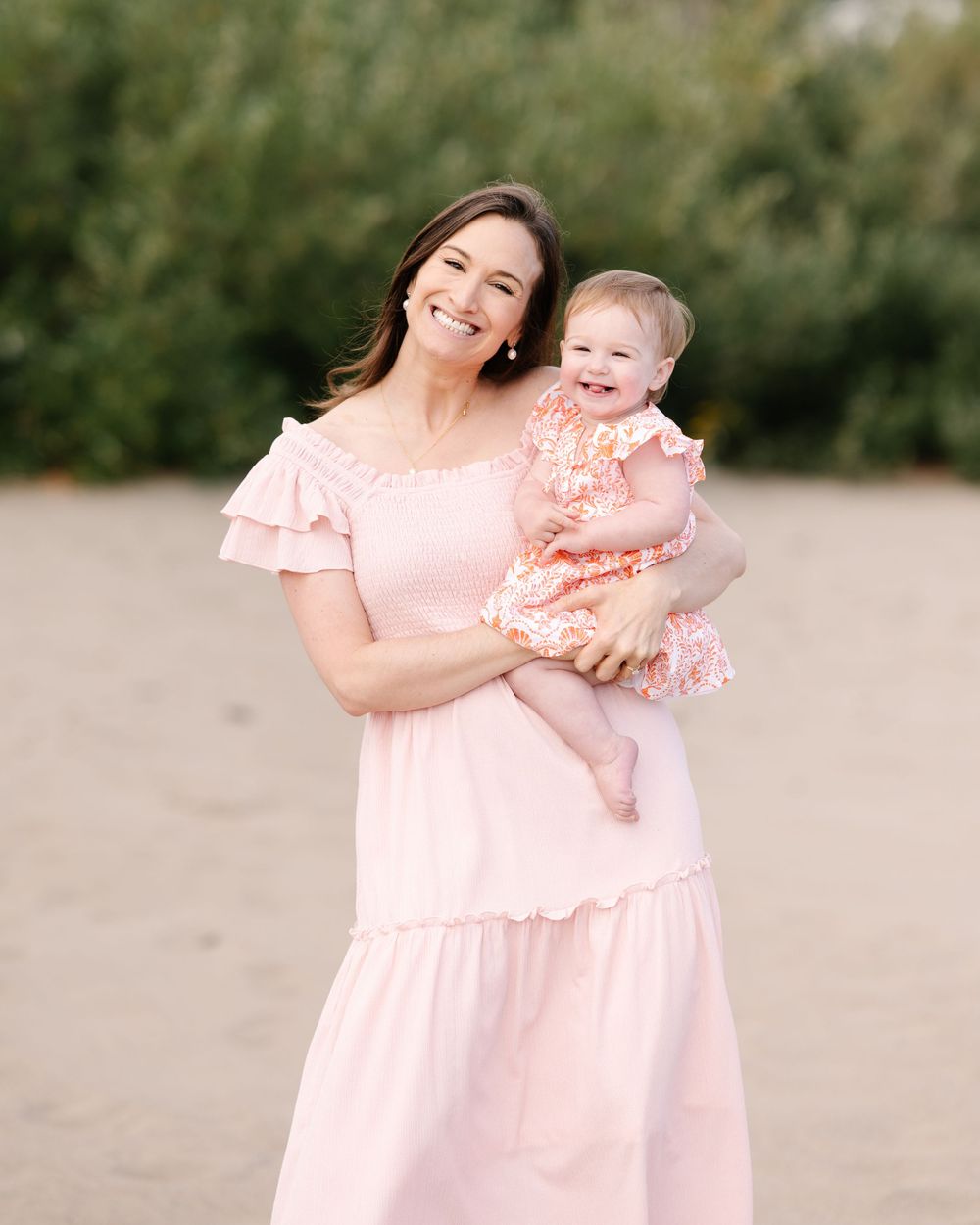 Mother in pink dress holding smiling baby girl on sandy beach.