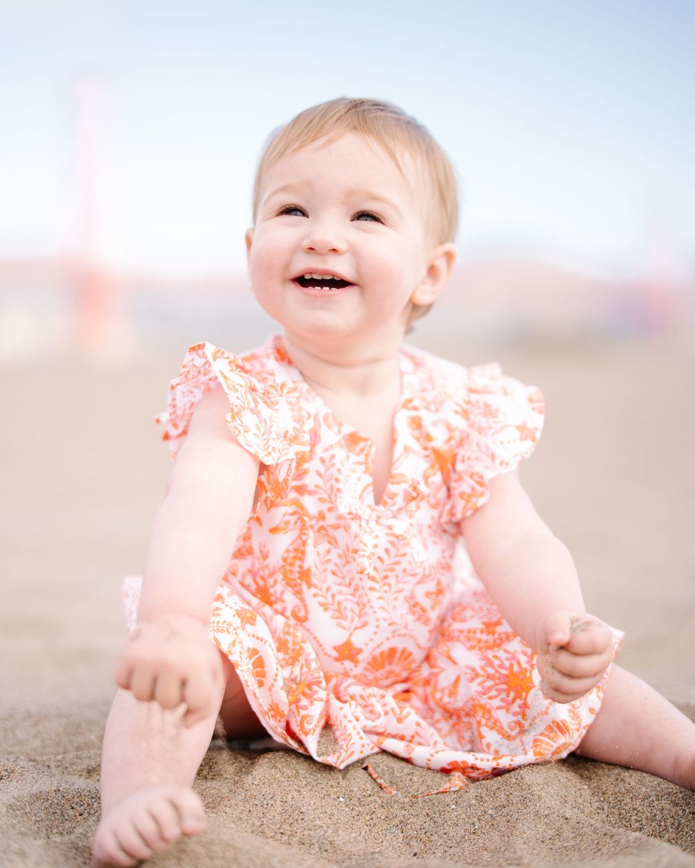 Smiling baby girl in orange floral romper sitting on sandy beach.