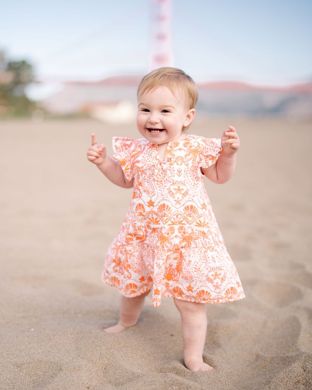 Happy baby girl in orange floral dress standing on sandy beach near Golden Gate Bridge, smiling and raising hands.
