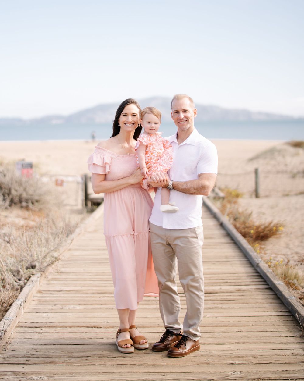 Family of three posing on a beachside boardwalk, mother in pink dress holding toddler, father in white shirt, mountains behind.