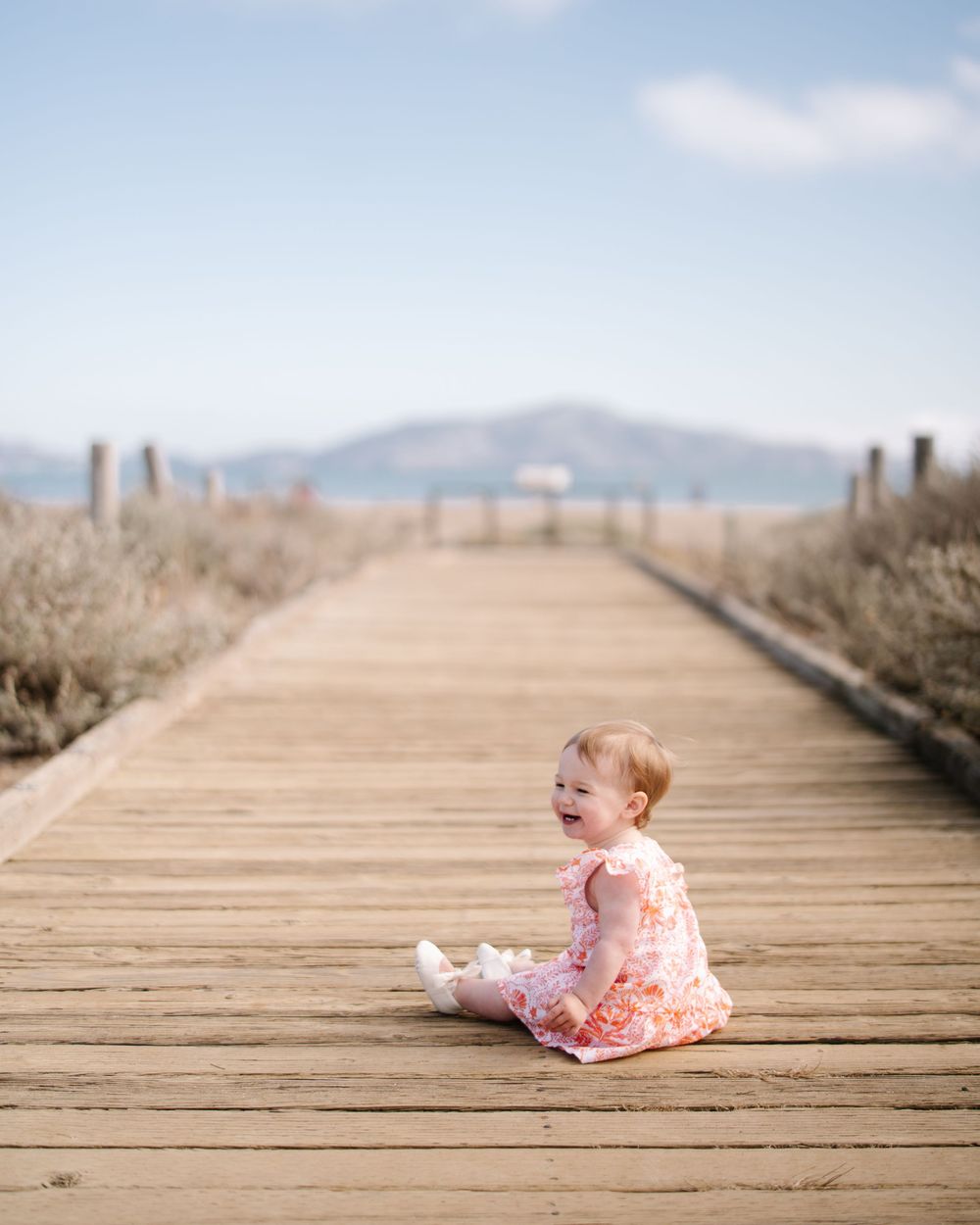 A smiling baby girl in a pink floral dress sits on a wooden boardwalk with mountains and blue sky behind her.