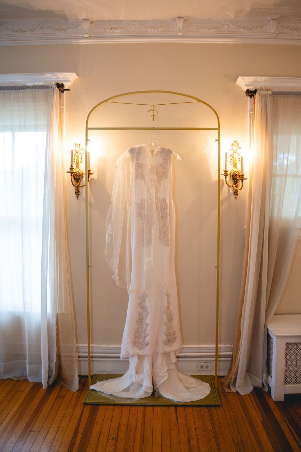 Elegant white wedding dress with lace details hanging on a gold rack flanked by ornate wall sconces.
