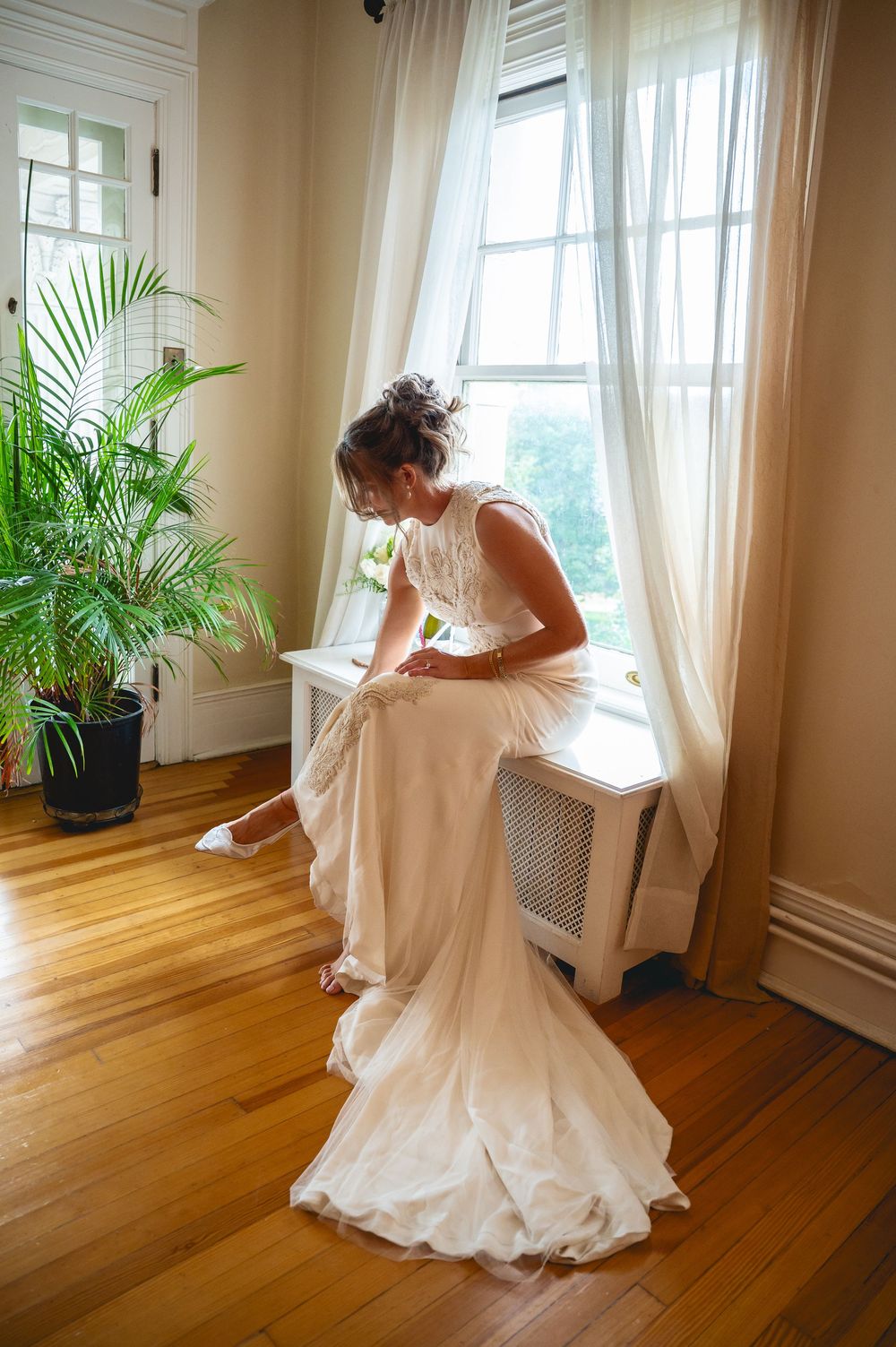 Bride in white wedding gown adjusting her shoe by a sunlit window with sheer curtains and a tropical plant nearby.