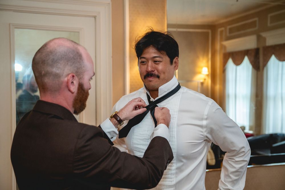A man in a white dress shirt gets his black tie adjusted by a friend in a brown suit at a wedding.