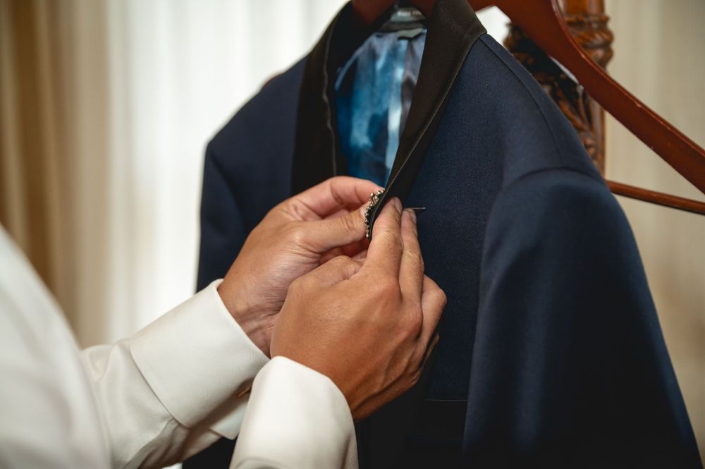 Hands adjusting a blue lapel pin on a navy suit jacket hanging on a wooden hanger.