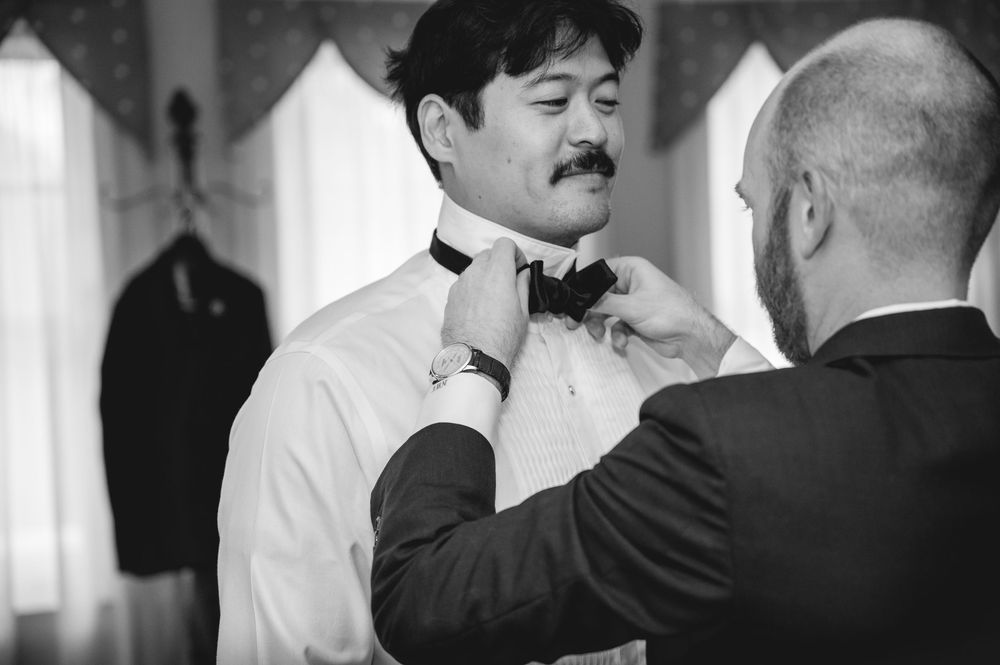 Groom getting bow tie adjusted by groomsman while preparing for wedding ceremony in black and white photo.