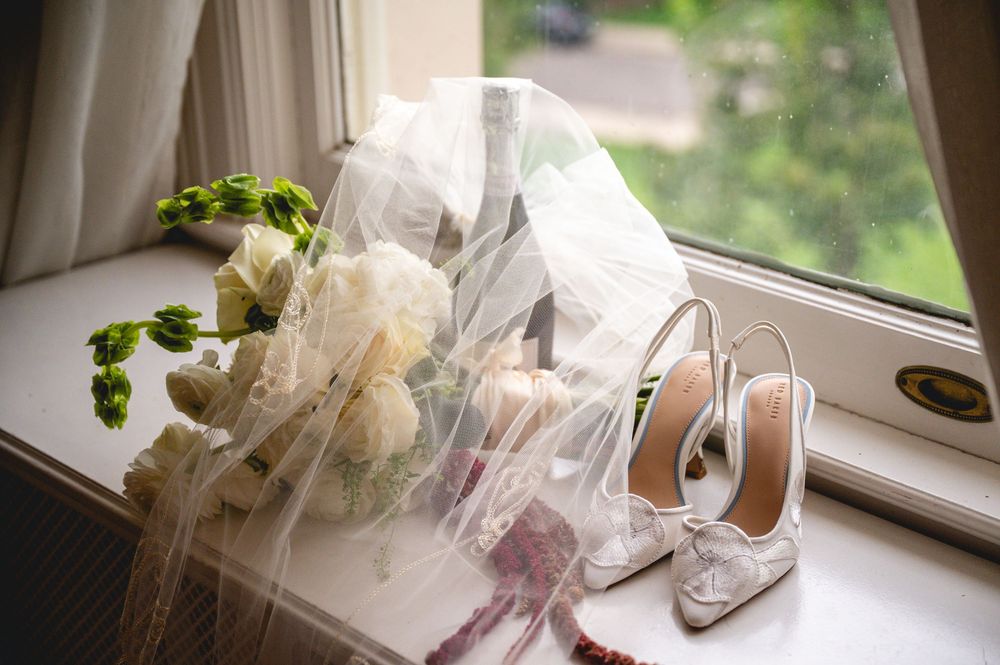 Bridal accessories on a windowsill featuring white lace heels, a bouquet of white roses, and a flowing veil.