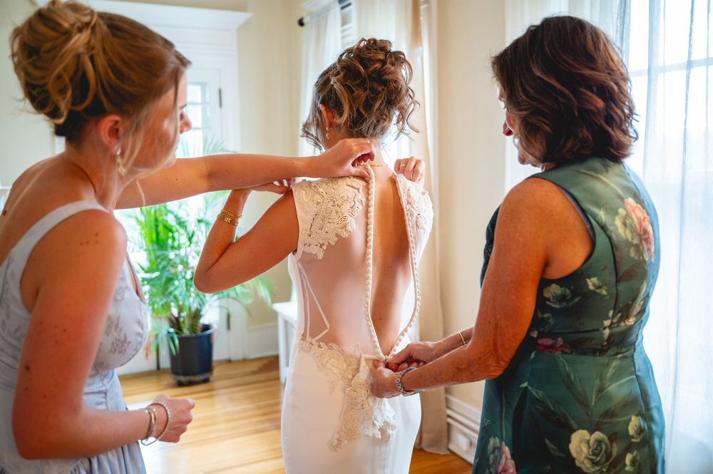 Two bridesmaids helping a bride button up her elegant lace wedding dress before the ceremony.