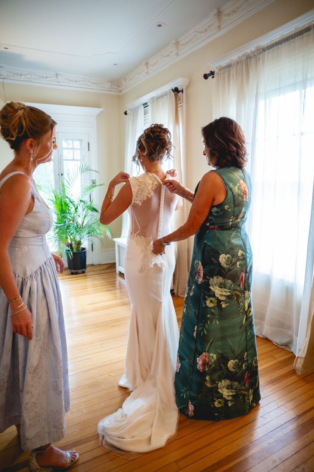Bride being helped into white lace wedding gown by two women in elegant dresses in a sunlit room.