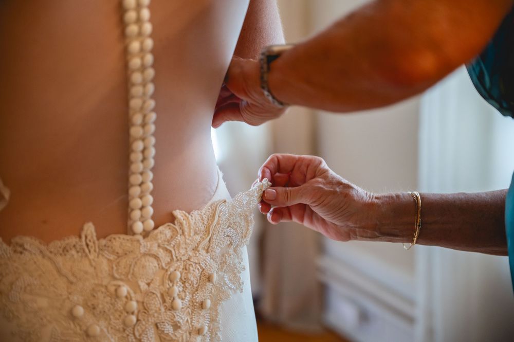 Close-up of hands fastening pearl buttons on a lace wedding dress during bridal preparation.