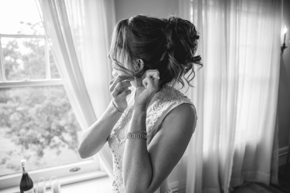 Bride putting on earrings in black and white photo by window in lace wedding dress.