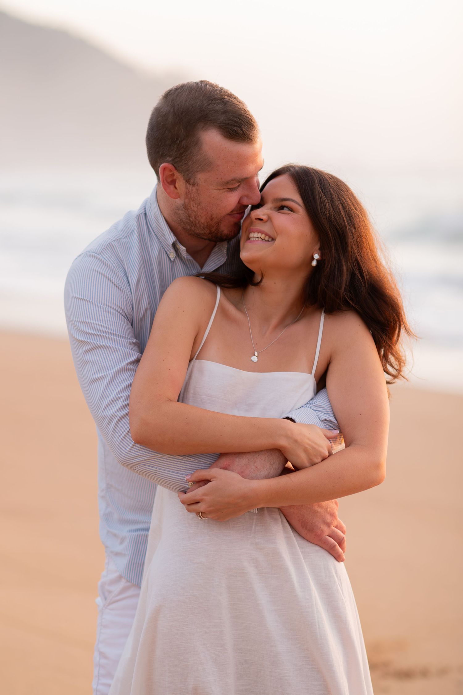 Romantic couple embracing on a sandy beach at sunset, woman in white dress smiling as man kisses her cheek.