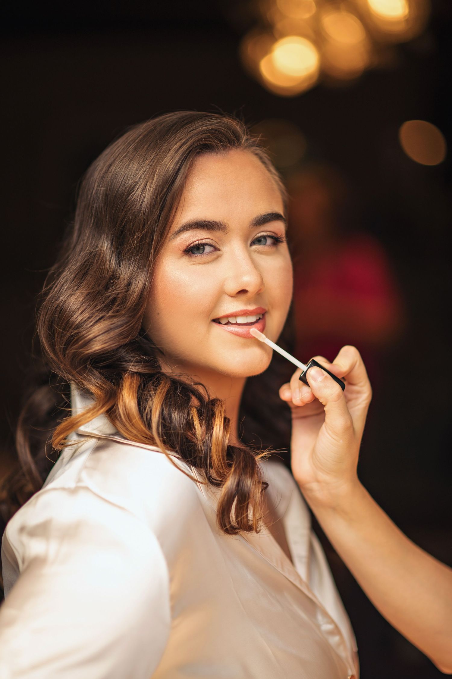 A young woman in a white robe gets lip gloss applied by a makeup artist, with warm bokeh lights in the background.