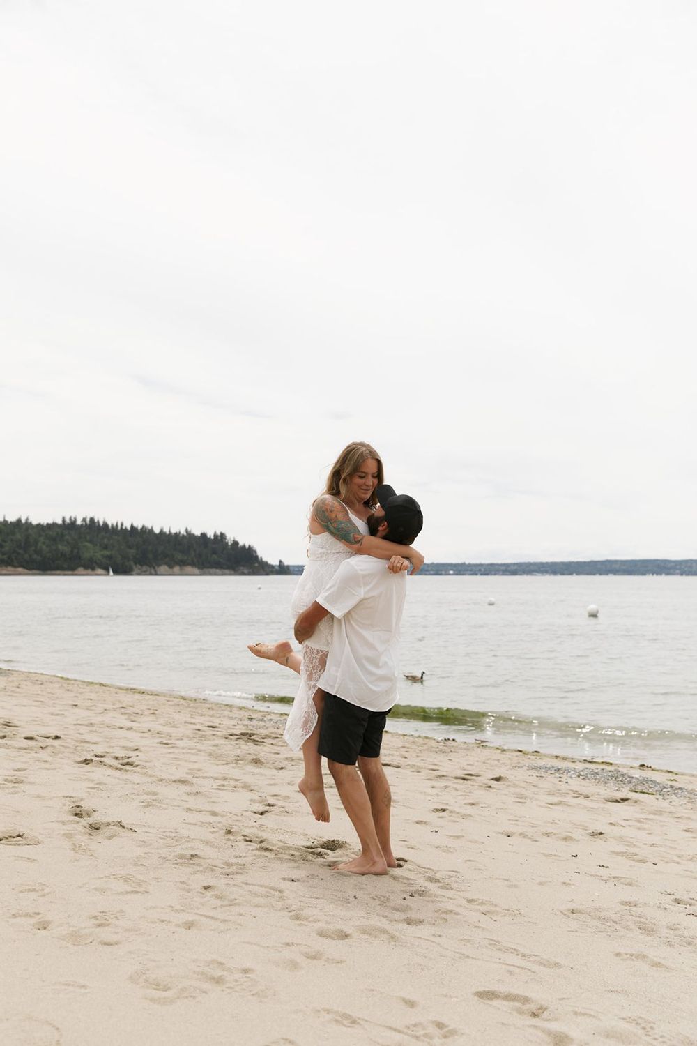 Romantic couple embracing on a sandy beach with calm ocean waters and evergreen trees in the background.