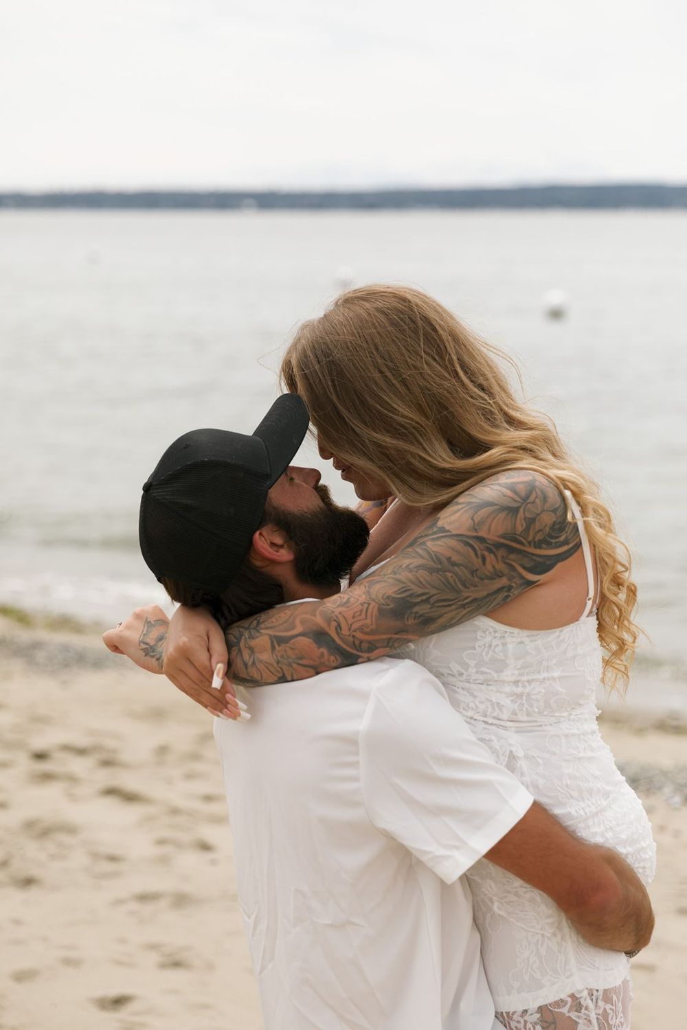Tattooed couple embracing and kissing on a sandy beach, woman in white lace dress, man in white shirt and black cap.