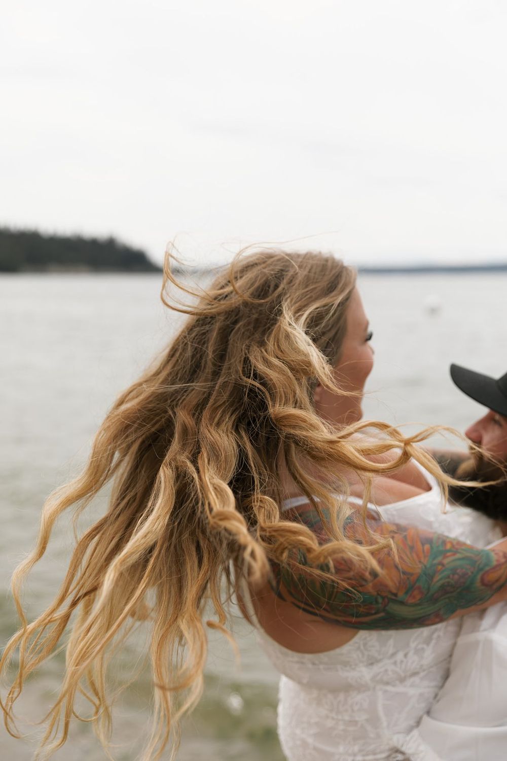 Woman with long blonde wavy hair blowing in the wind being embraced by a tattooed partner near the ocean.